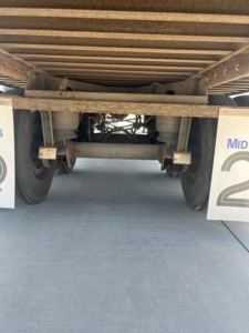 Underneath a utility van trailer showing rear axle assembly, leaf springs, and mud flaps with partial 'Mid' signage. Rust and metal frame visible.