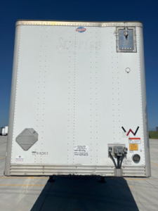 Rear view of a white utility van trailer showing a small door, clearance light, and connection hardware with riveted panels and a diamond-shaped vent; markings and labels near bottom.