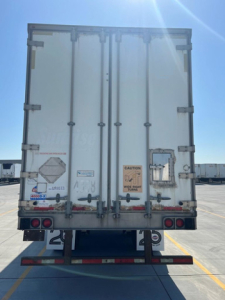 Rear view of a white cargo trailer with double doors, warning stickers, and taillights in a parking lot.