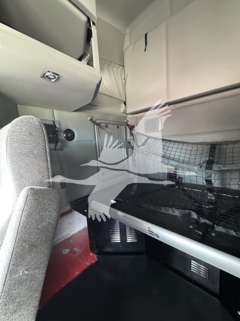 Kenworth T680 cab interior showing sleeper area with storage cabinets, mesh cargo net, and control panel; cab walls light gray.