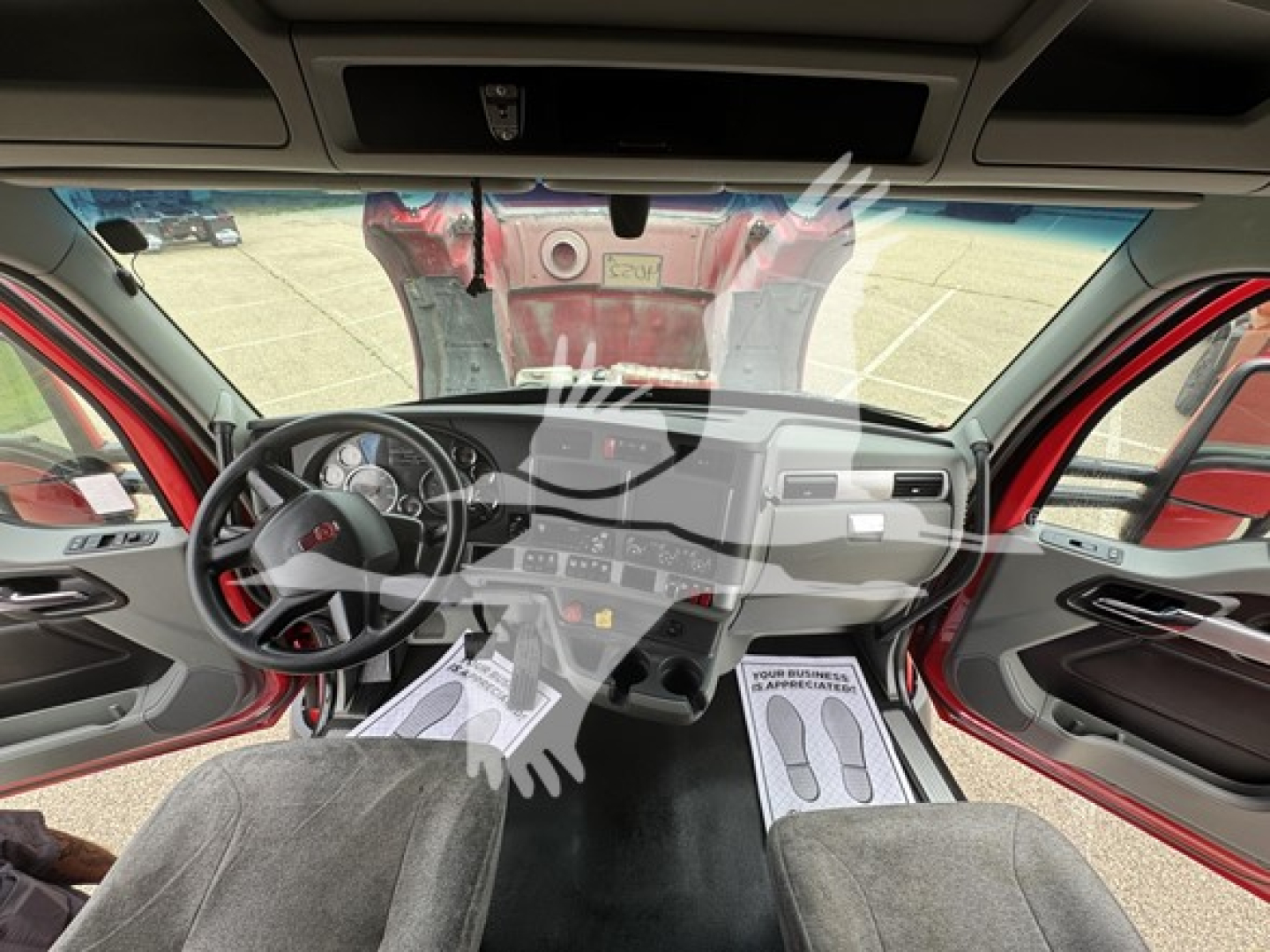 Kenworth T680 cab interior view with steering wheel, dashboard, and driver seat visible; windshield showing outside parking lot and a red truck behind.