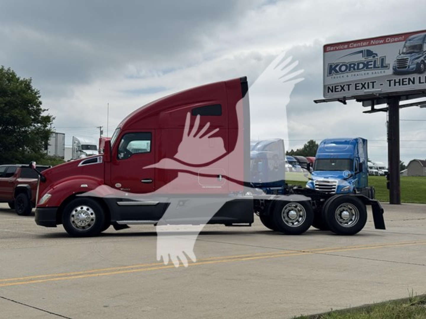 Kenworth T680 tractor in a parking lot, red cab, side view, with a blue truck in the background and a Kordell sign in the distance.