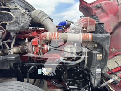 Kenworth T680 engine bay with intake piping, hoses, and coolant reservoir visible; red cab and black chassis components beneath.