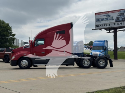 Kenworth T680 tractor in a parking lot, red cab, side view, with a blue truck in the background and a Kordell sign in the distance.