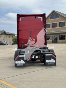 Back of red Kenworth trailer chassis with fifth wheel and rear lights, two Kordell mud flaps, and asphalt lot in background.