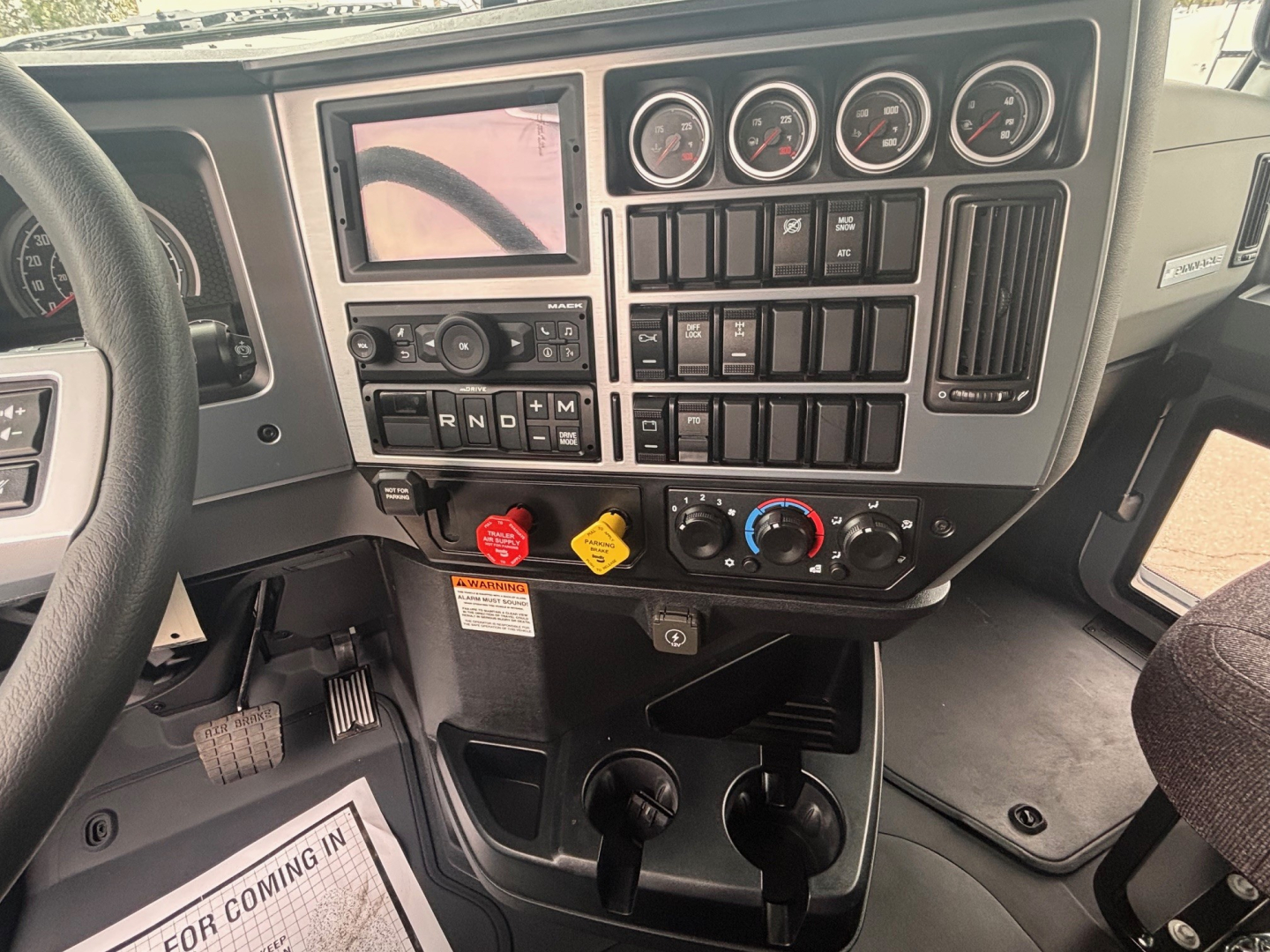 Mack PI64 dash panel showing instrument cluster, switches, and gear selector area with coolant and warning labels. Includes steering wheel left and cup holders under center console.