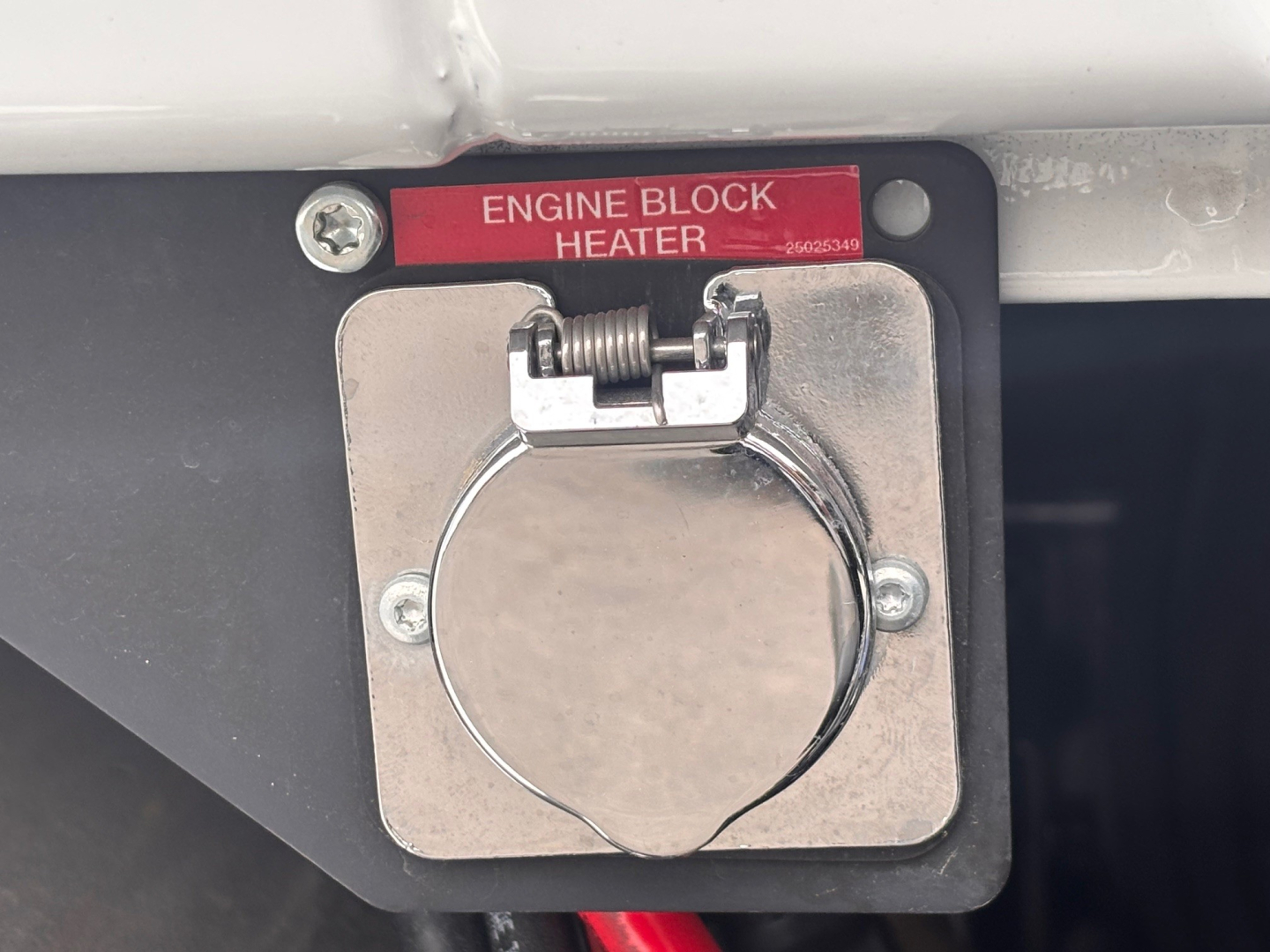 Diesel truck engine block heater plate with red label reading ENGINE BLOCK HEATER and circular cap, mounted on firewall area of Mack PI64 truck
