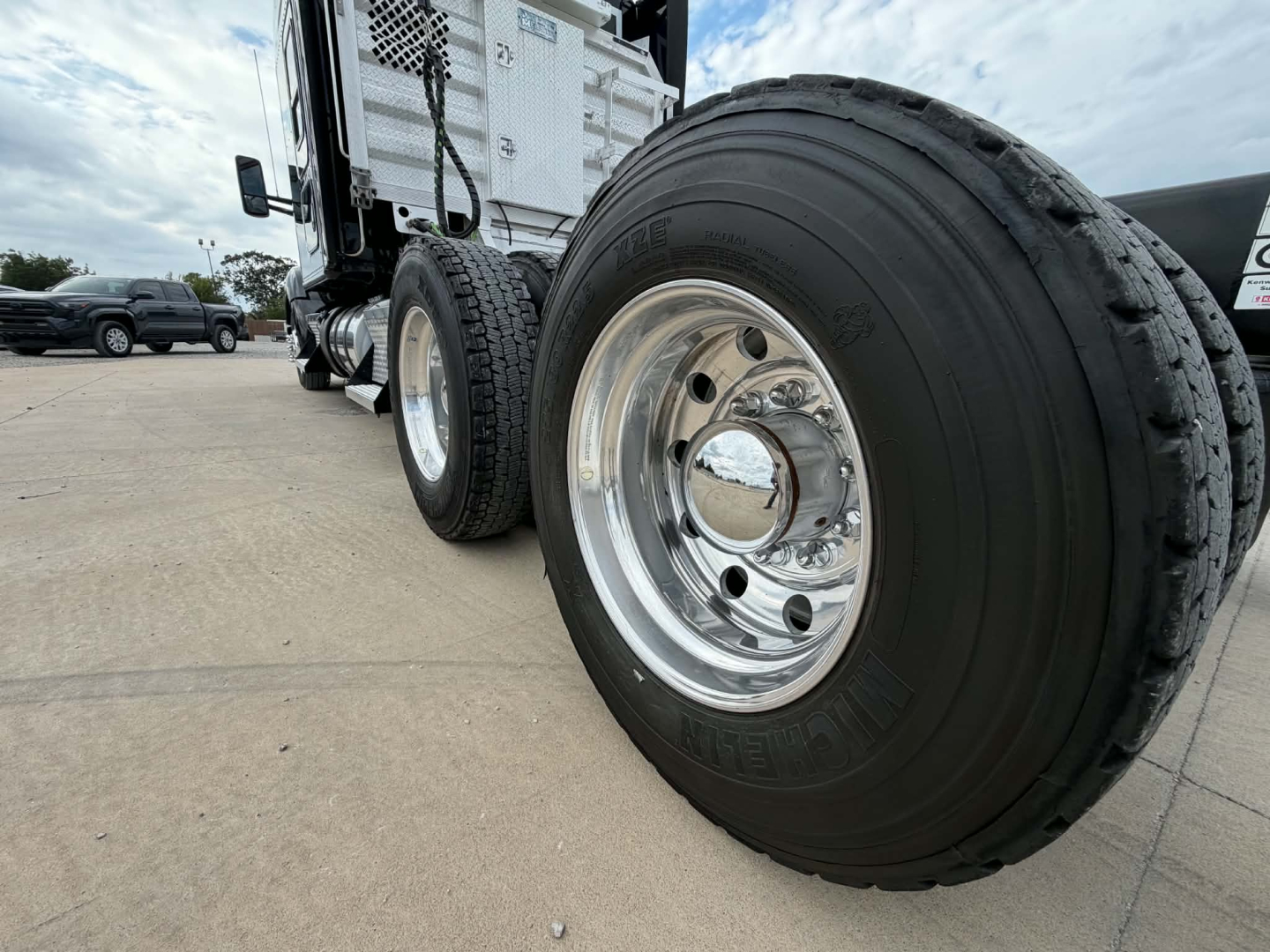 Dual rear wheels and tires of a Kenworth truck in the foreground, with chrome rims and heavy-duty hub; part of the cab visible in background.