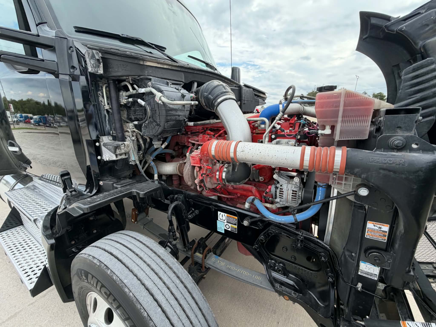 Kenworth T680 engine bay view showing red engine and hoses with intake piping, front wheel visible and exposed chassis.
