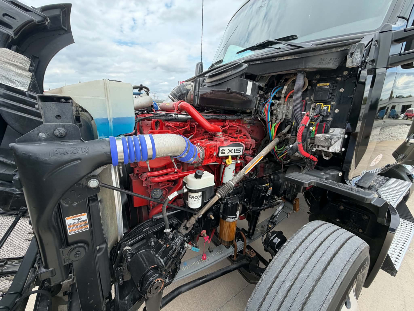 Kenworth T680 engine bay view showing red Cummins X15 engine and surrounding components with hoses, wiring, and coolant reservoir.