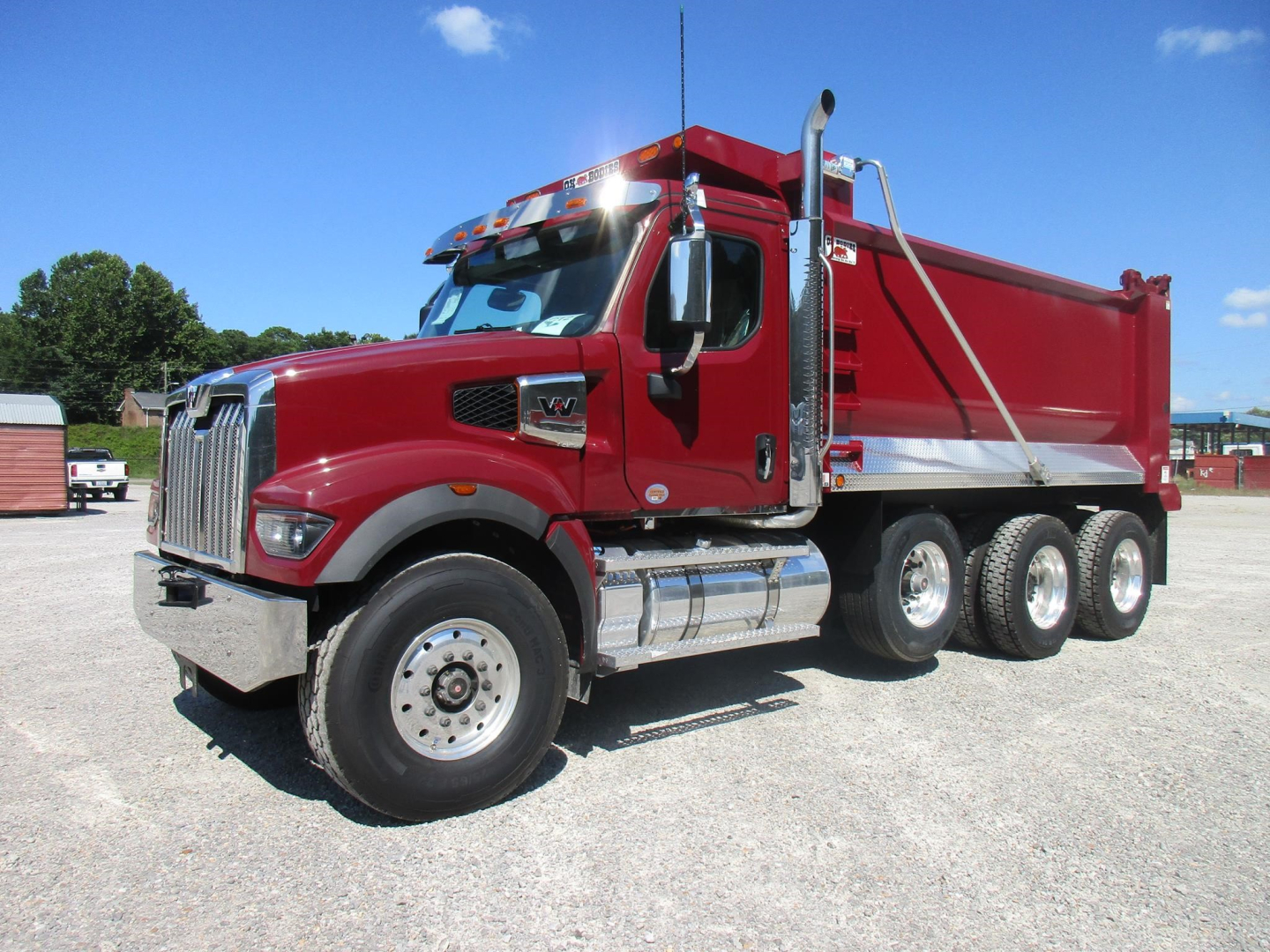 Dump truck in red shows Western Star 49X with chrome fuel tanks and triple rear axles, side view highlighting dump bed and cab features.
