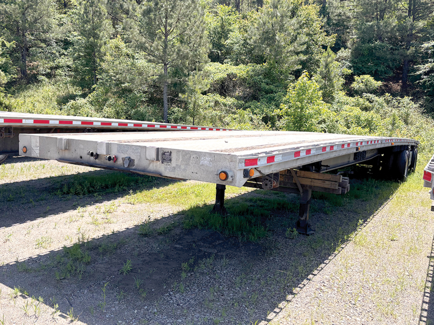 Flatbed trailer in side view showing long aluminum deck, red-white reflective tape, support legs retracted, and wooded background.