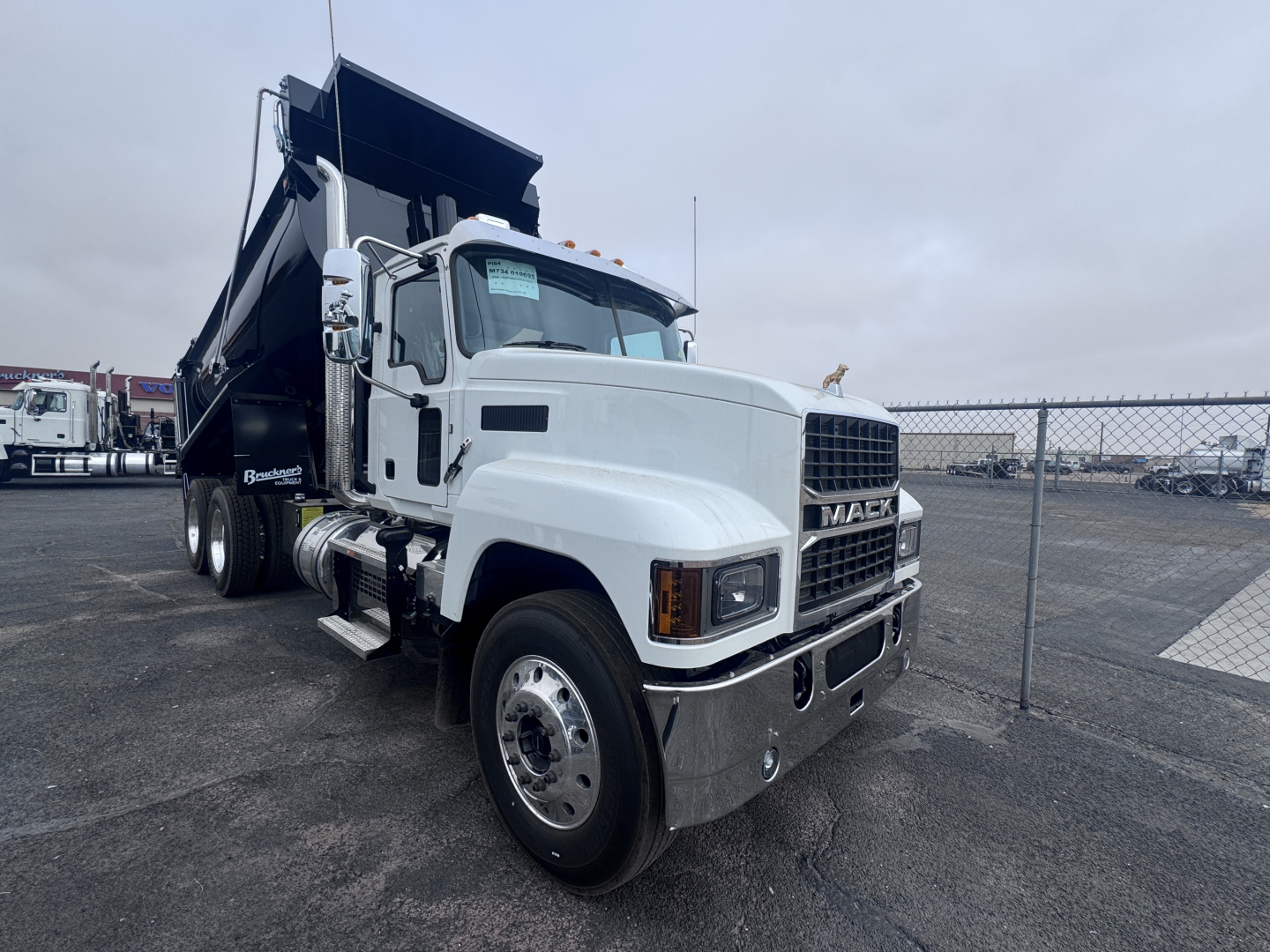 Truck front-left view showing a white Mack PI64 dump truck with open-box bed, chrome bumper, dual rear wheels, and a chain-link fence in the background.