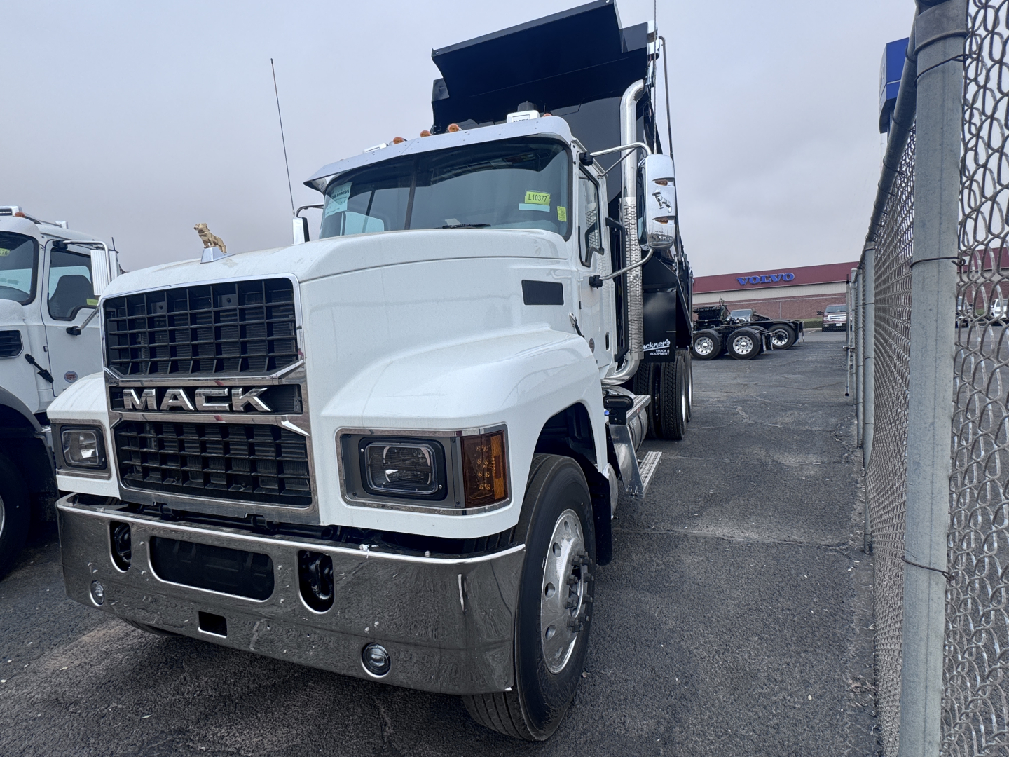 Mack PI64 dump truck in a lot showing front grille with MACK lettering, large chrome bumper, headlights, and dump bed raised. Visible text includes MACK on grille and truck body styling.