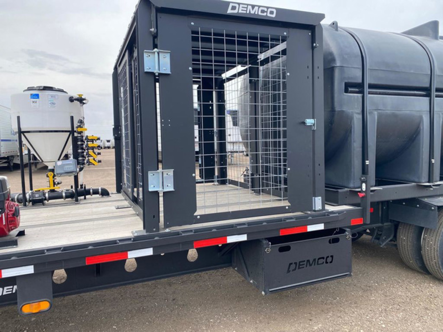Demco liquid tender trailer with a metal cage gate and side access door, mounted on a flatbed trailer with Demco branding visible on the equipment and bumper.