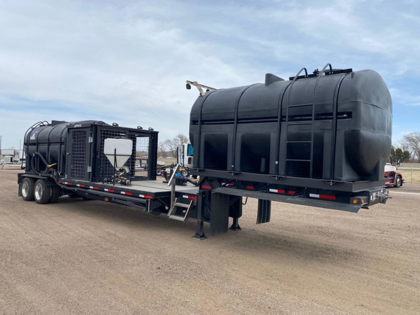 Trailer showing a large black Demco Liquid Tender saddle tank mounted on a flatbed trailer with twin rear axles and discharge hose assembly.