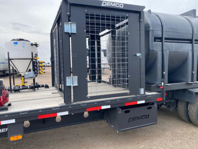 Demco liquid tender trailer with a metal cage gate and side access door, mounted on a flatbed trailer with Demco branding visible on the equipment and bumper.