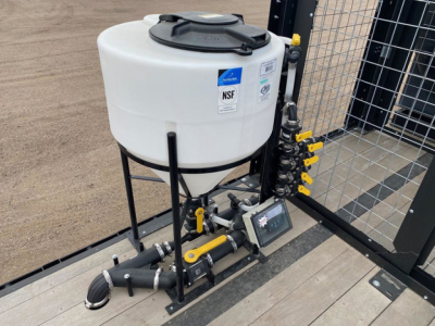 Demco Liquid Tender trailer equipment with white vertical cylindrical tank on a black metal frame, mounted hoses, fittings, and a control panel on a trailer bed.