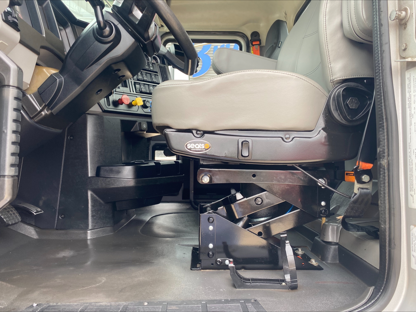 Cab interior of a 2019 Mack PI64T daycab showing driver seat and control panel with steering wheel, dashboard switches, and gear/mechanism under seat.