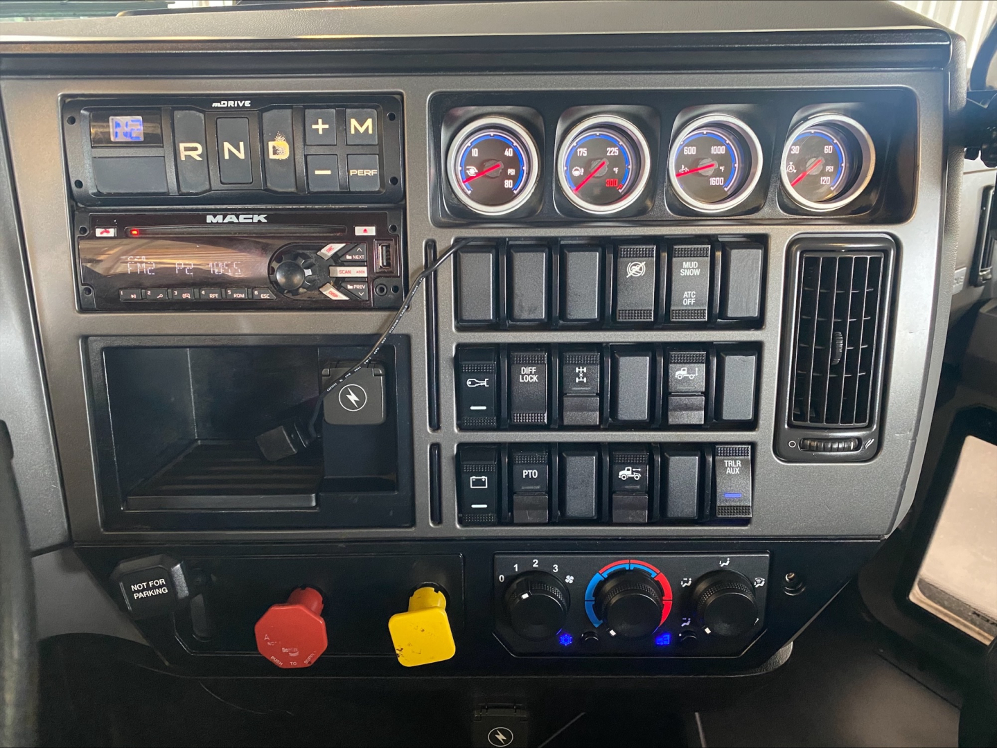 Truck dashboard of a 2019 Mack PI64T showing gear selector, three gauges, various switches, and climate controls.