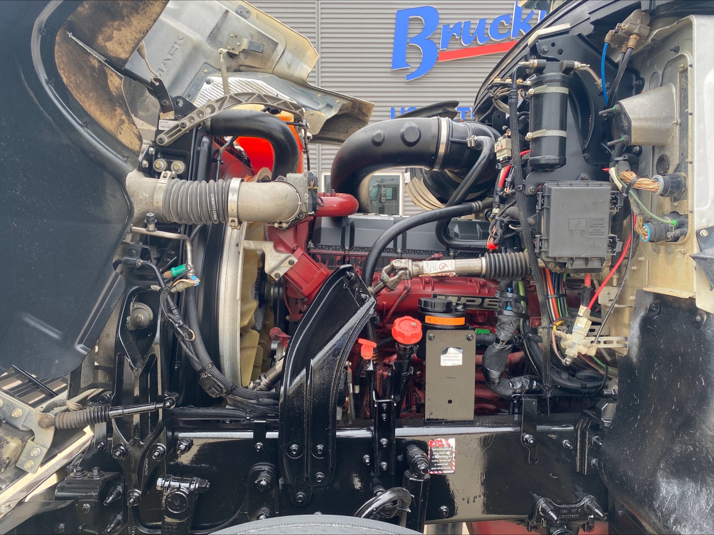 Mack PI64T engine bay with visible hoses, wiring, and components including air intake, coolant reservoir, and hydraulic lines.
