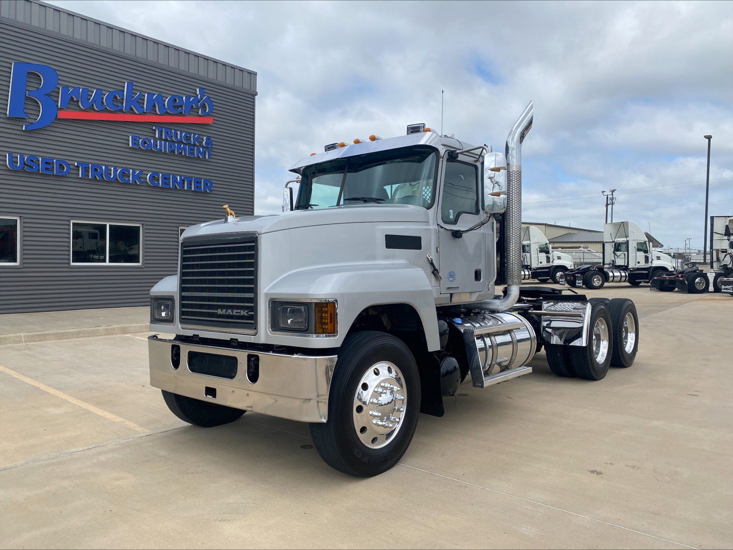 Mack PI64T conventional daycab truck shown from front-left angle with gray cab and chrome bumper, large exhaust stacks, and dual rear axles in a dealership lot.