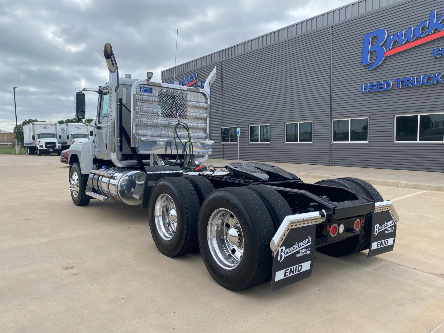 Truck chassis rear view showing dual rear axles, mud flaps, chrome fuel tanks, and silver diamond plate cab splash guard on a 2019 Mack PI64T conventional daycab.