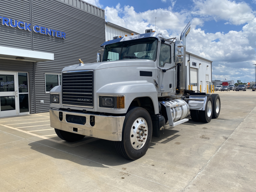 Mack PI64T conventional daycab truck in front of a vehicle center, showing front grille, white cab, dual rear axles, and chrome fuel tanks.