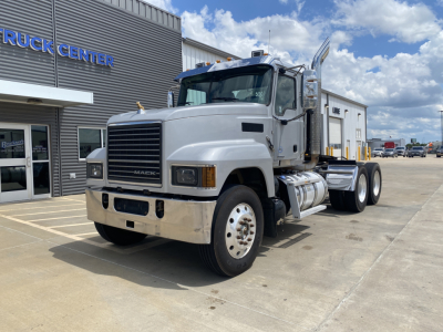 Mack PI64T conventional daycab truck in front of a vehicle center, showing front grille, white cab, dual rear axles, and chrome fuel tanks.