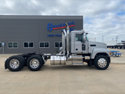Mack PI64T Conventional Daycab truck side view showing fuel tanks, chrome exhaust stacks, and dual rear axles in front of a dealership building.