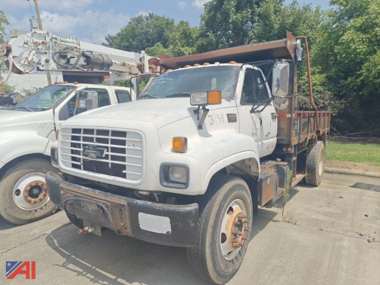 Dump truck with white cab and flatbed, front grille with Chevrolet emblem, orange warning lights, and a hydraulic hose setup; visible rust on bumper and wheel hubs.