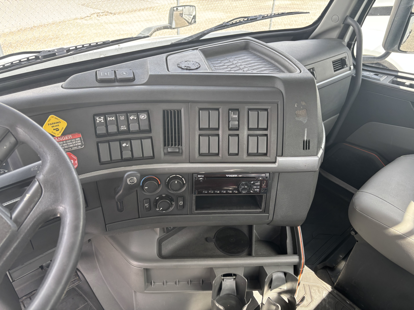 Volvo VNL dashboard and center console with control switches and radio; steering wheel visible on left, gray plastic interior, cup holder and storage compartments.