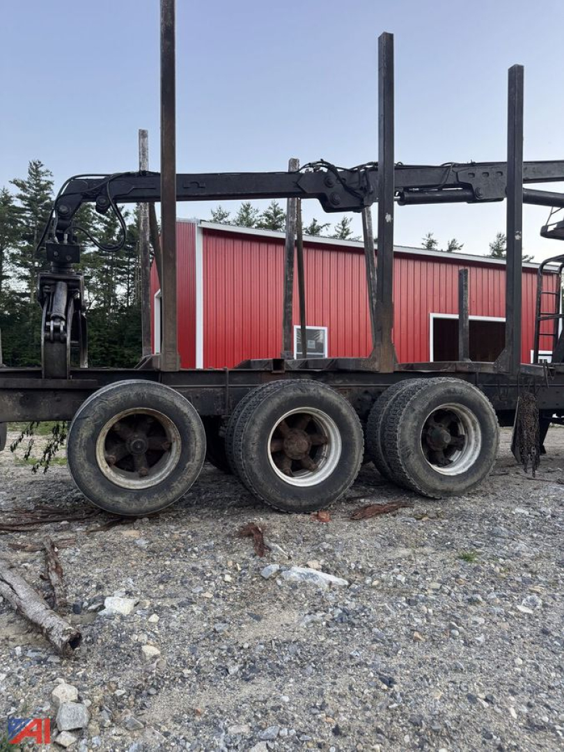 Log trailer frame with four wheels and a hydraulic arm, black paint, metal rails, and a red building in the background.