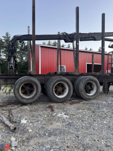 Log trailer frame with four wheels and a hydraulic arm, black paint, metal rails, and a red building in the background.
