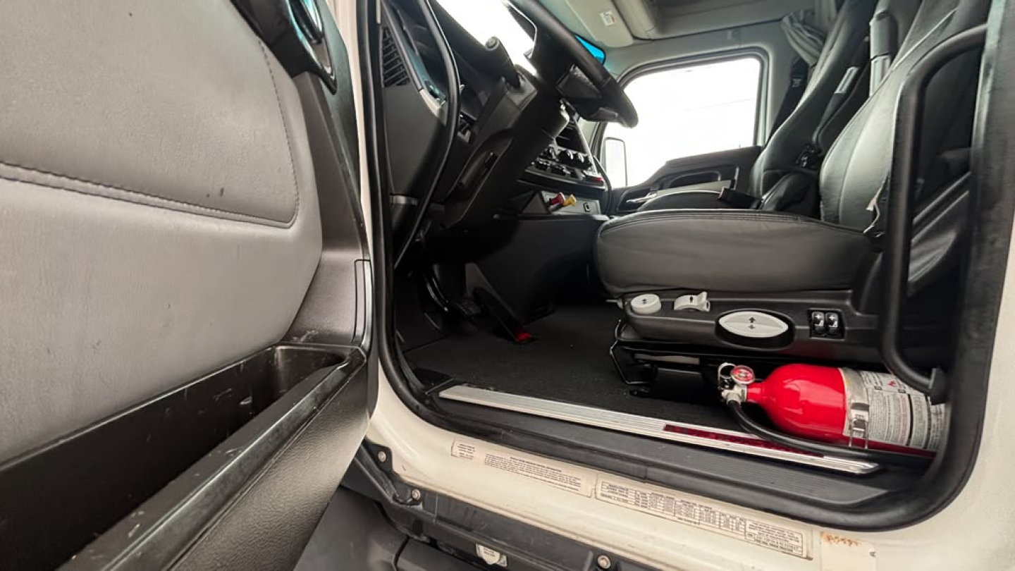 Cabin of a 2024 Kenworth T680 tractor-trailer showing front seat, dashboard, and interior with a red fire extinguisher visible near the floor.