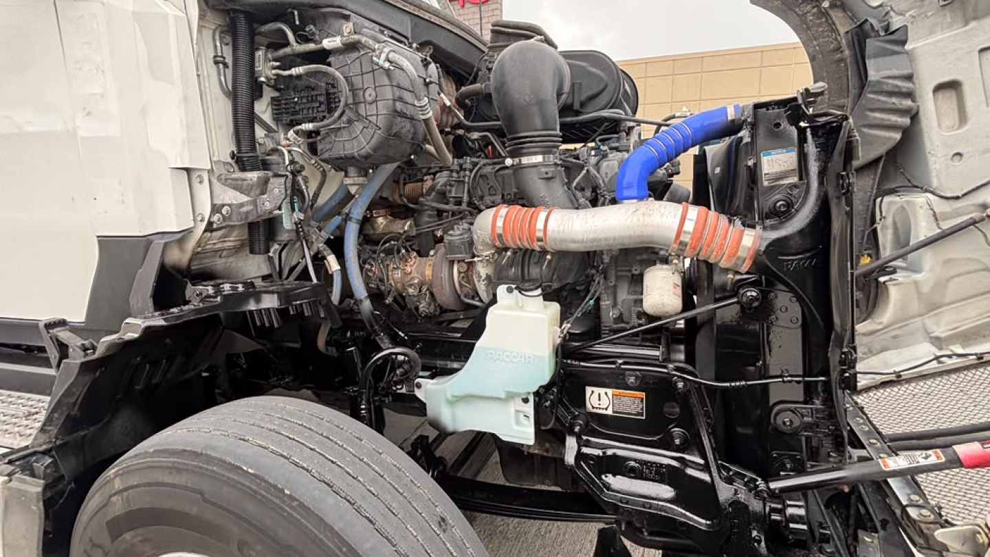 Kenworth T680 engine bay view showing cooling reservoir, hoses, turbo intake, and various engine components in a truck. Visible label areas and wiring harnesses.