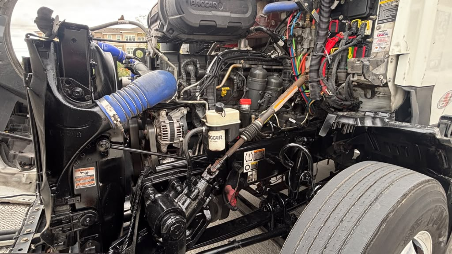 Kenworth T680 engine bay with exposed components including hoses, wiring, and fluid reservoirs. Visible labels include oil and coolant caps, belts, and a blue silicone hose.
