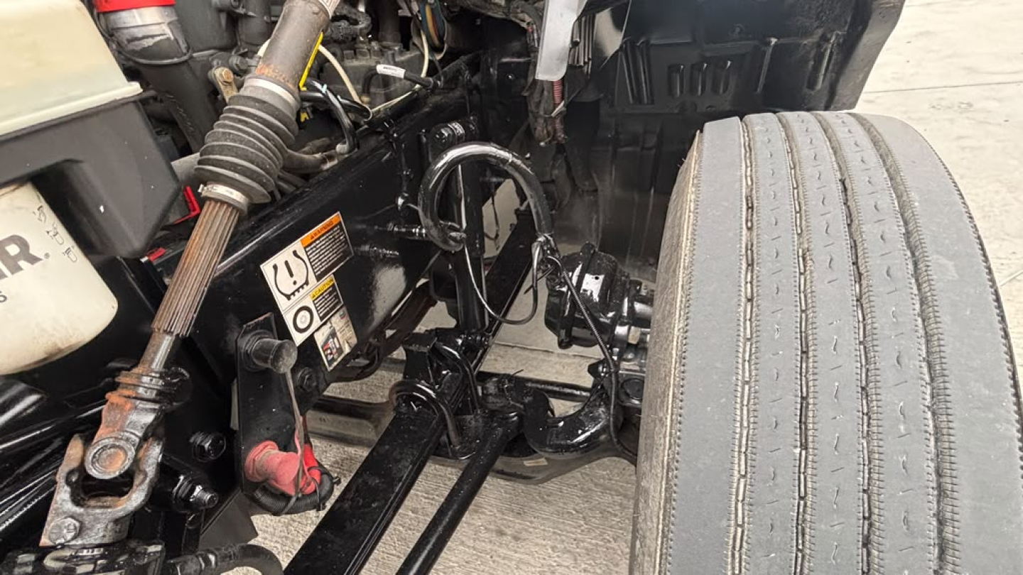 Kenworth T680 engine bay area showing front suspension and steering components, visible hoses and wiring, and a single front tire with tread.