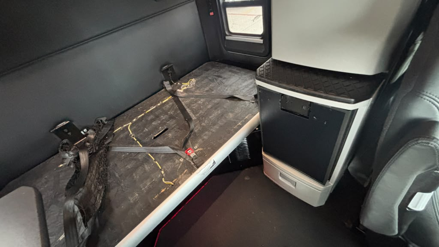 Kenworth T680 sleeper cab interior showing rear bunk area with seat/bench and window, worn surface with seat belts and storage cabinet.