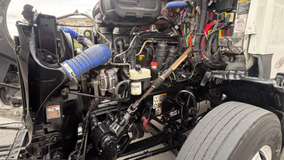 Kenworth T680 engine bay with exposed components including hoses, wiring, and fluid reservoirs. Visible labels include oil and coolant caps, belts, and a blue silicone hose.