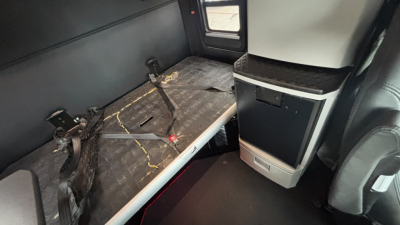 Kenworth T680 sleeper cab interior showing rear bunk area with seat/bench and window, worn surface with seat belts and storage cabinet.