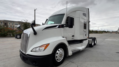 Truck side view showing 2024 Kenworth T680 conventional sleeper, white exterior, dual rear axles, large front grille, aerodynamic mirrors.