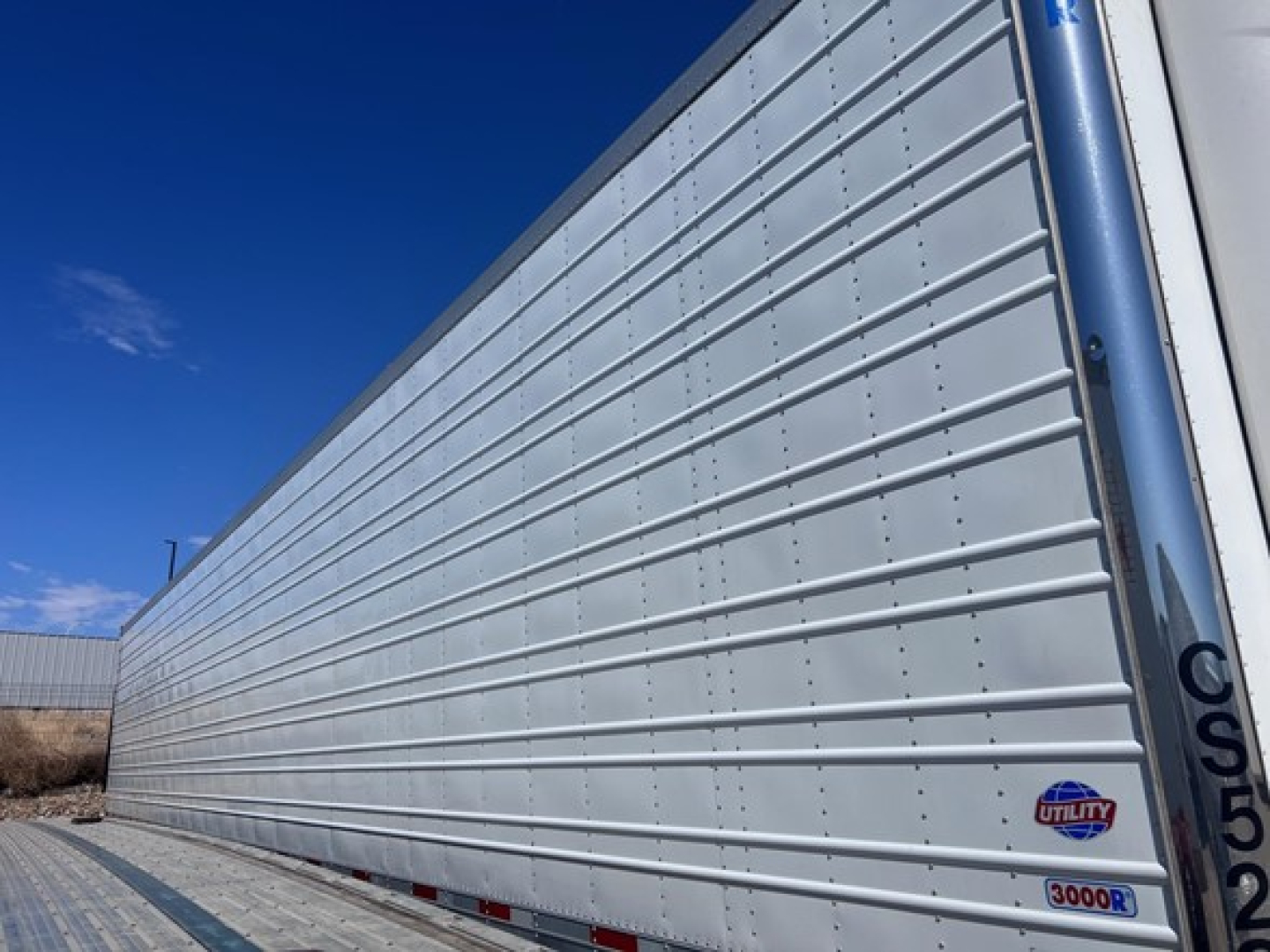 Long silver Utility 3000R reefer trailer side with vertical corrugation and small circular Utility logo near front, showing chrome riveted panels.