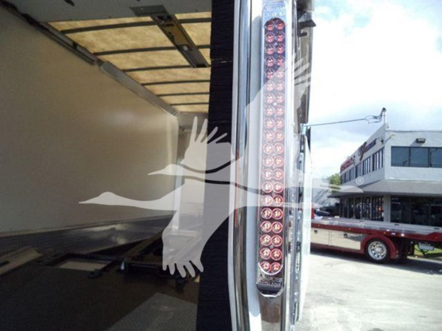 Mack MD6 truck taillight area with vertical LED strip and open trailer; reflection of building and other vehicles visible.