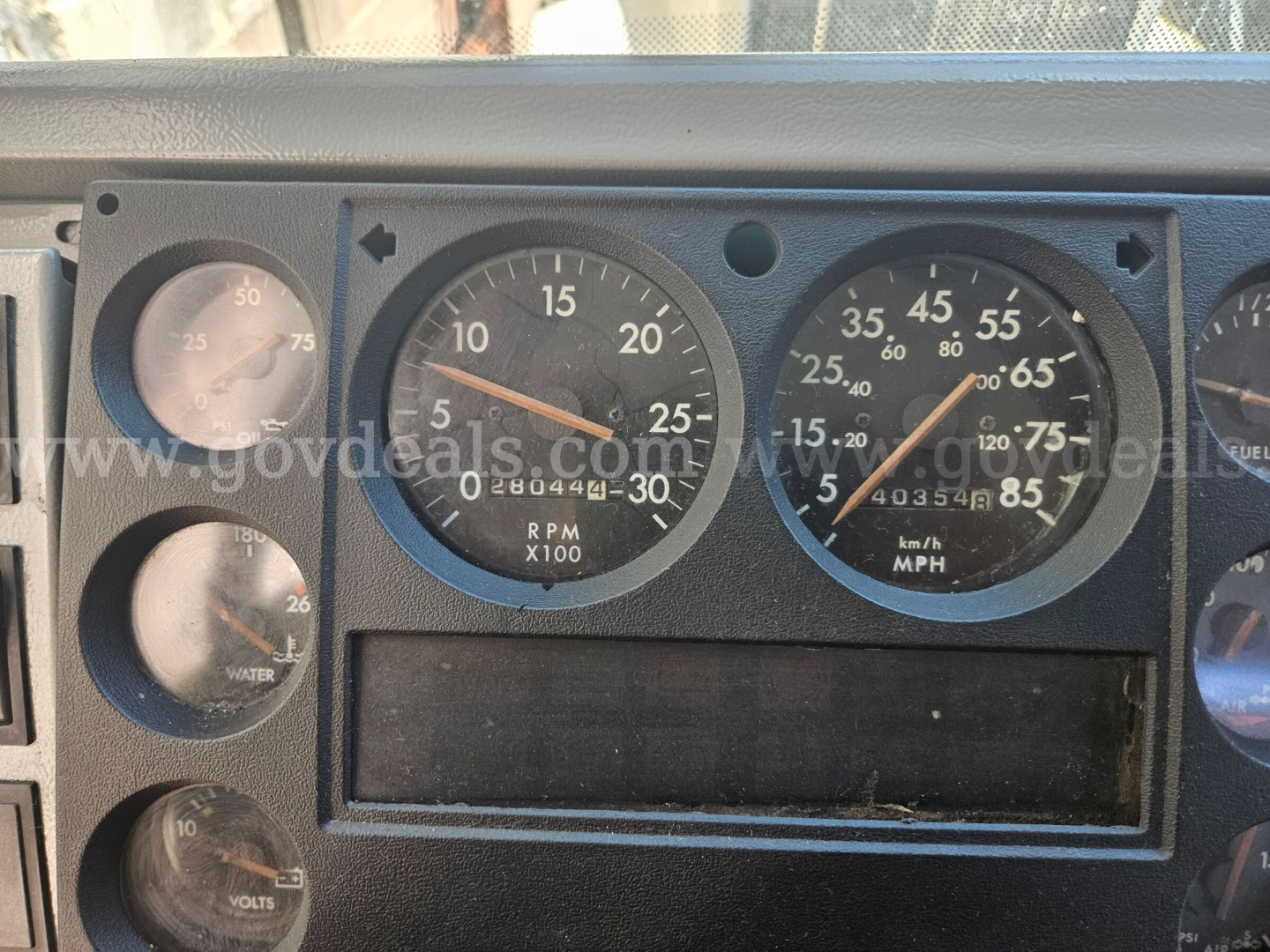 Dashboard cluster from a Freightliner cab showing rpm gauge X100, speedometer mph, various dials, and blank digital readout. Visible markings include RPM 0-30, SPEED 0-85, and smaller gauges with numbers.