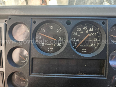 Dashboard cluster from a Freightliner cab showing rpm gauge X100, speedometer mph, various dials, and blank digital readout. Visible markings include RPM 0-30, SPEED 0-85, and smaller gauges with numbers.