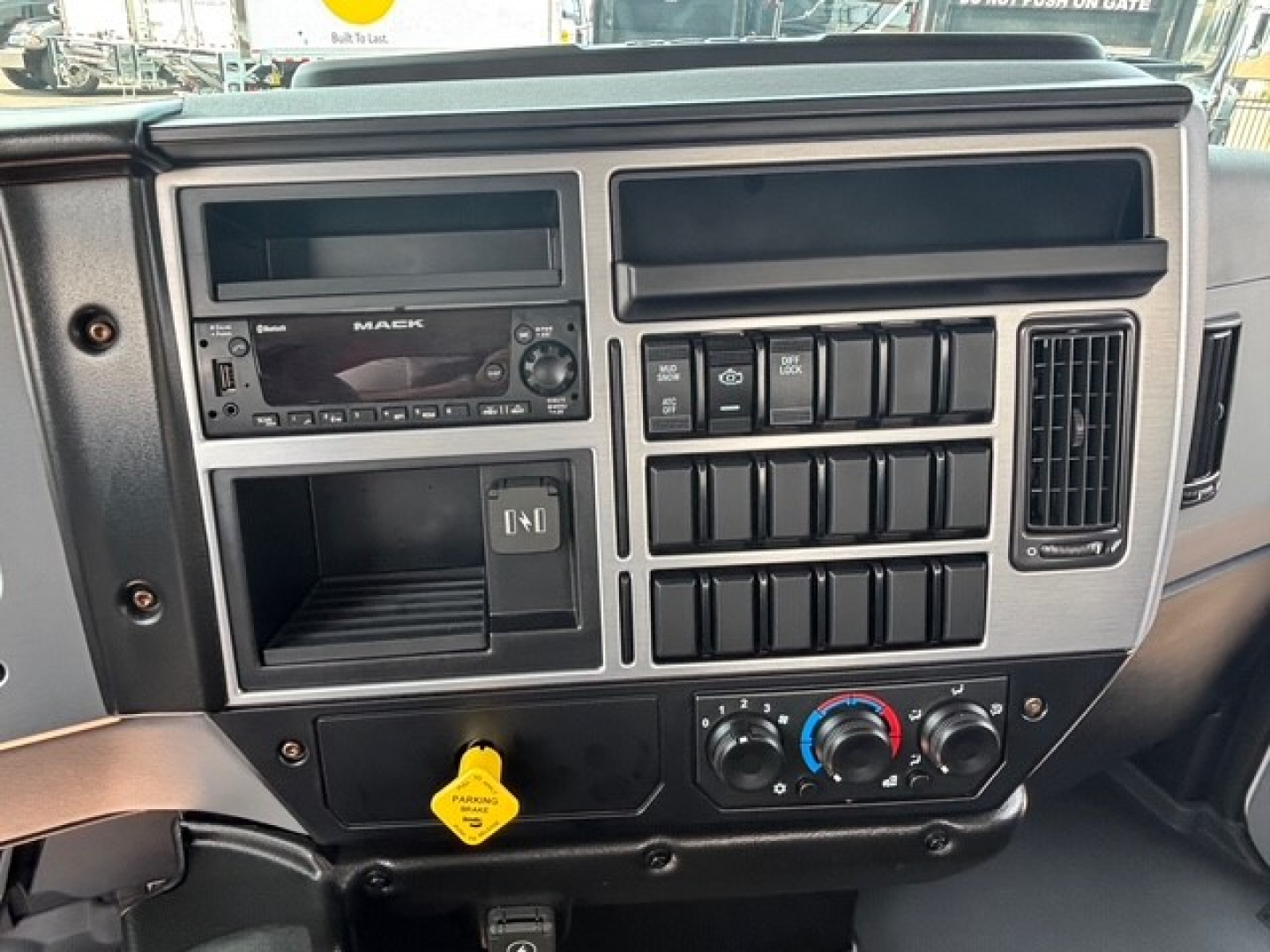 Dashboard of a 2026 Mack MD7 cab; visible features include a Mack radio unit, multiple control switches, circular climate controls, and a yellow parking brake knob.