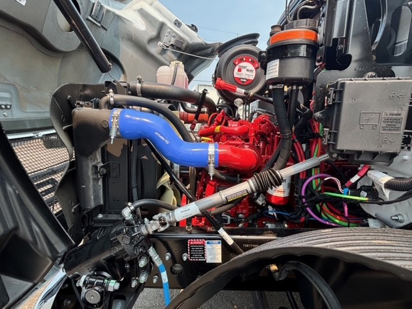 Mack MD7 engine bay showing blue and red hoses, gauge, and wiring harnesses in a flatbed truck engine compartment.