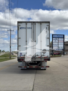 Rear view of a white utility trailer with River Valley branding and a West Ridge Business Center sign, showing taillights, bumper, and rear doors.
