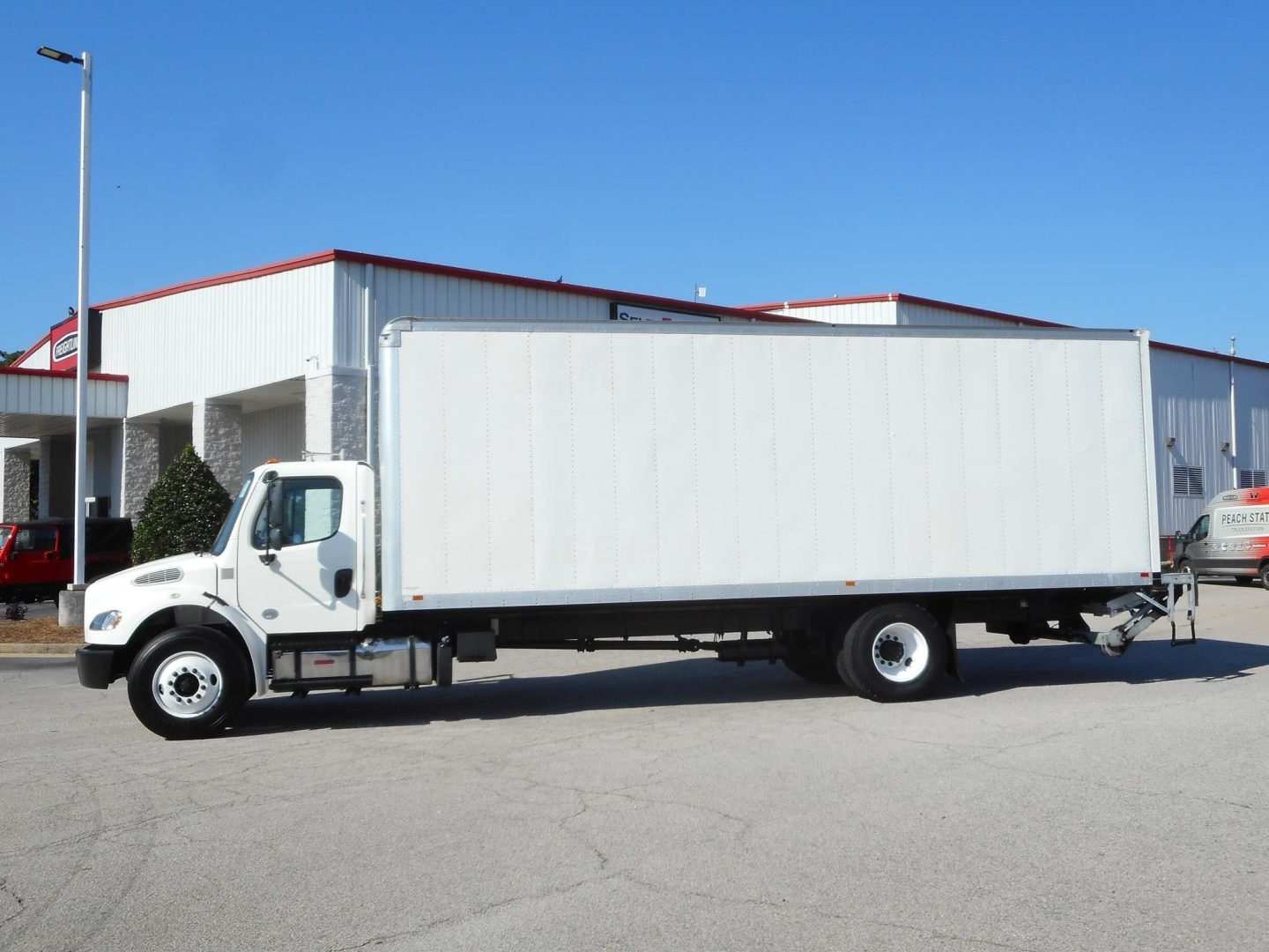 Medium-duty Freightliner M2 106 box truck side view with white cargo box, single rear axle, cab at left, dealership lot in background